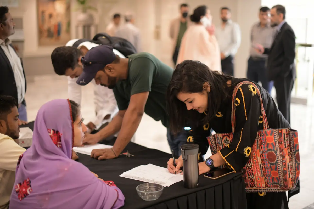 People gather around a registration table at an indoor event in Pakistan, some filling out paperwork and others conversing in the background.
